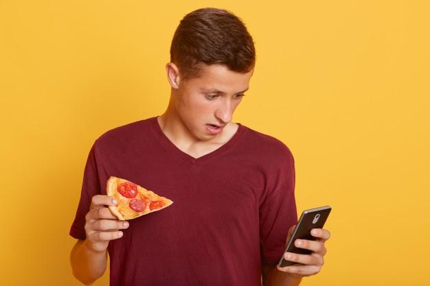 Surpreendente bonitão, segurando a fatia de pizza e telefone saborosos, verificando a rede social enquanto come um lanche, vê notícias de frenagem