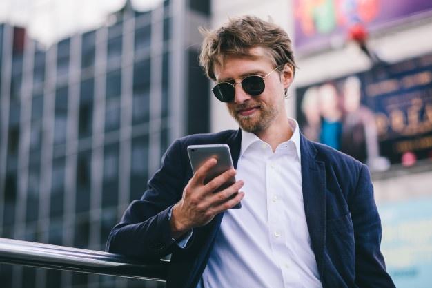 Um jovem rapaz com cabelos loiros em óculos de sol olha para o seu celular enquanto estava ao ar livre num dia de verão